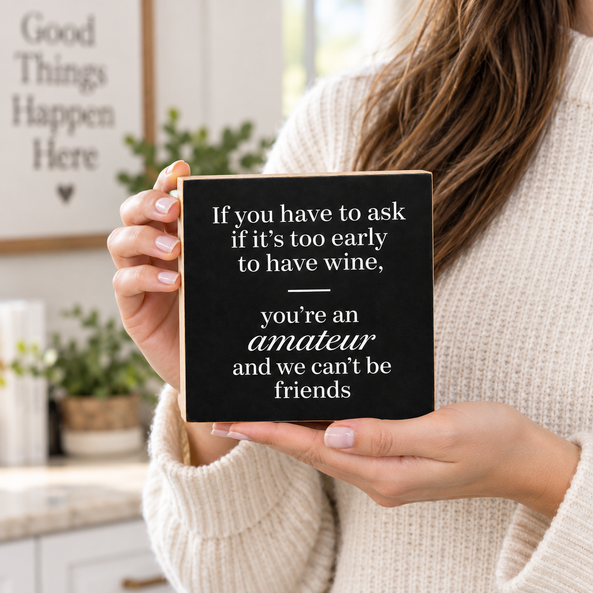 A person in a cream knit sweater holds a square black sign with white text that reads, 'If you have to ask if it's too early to have wine, you're an amateur and we can't be friends.' The background shows a softly blurred indoor setting with plants and a framed sign.