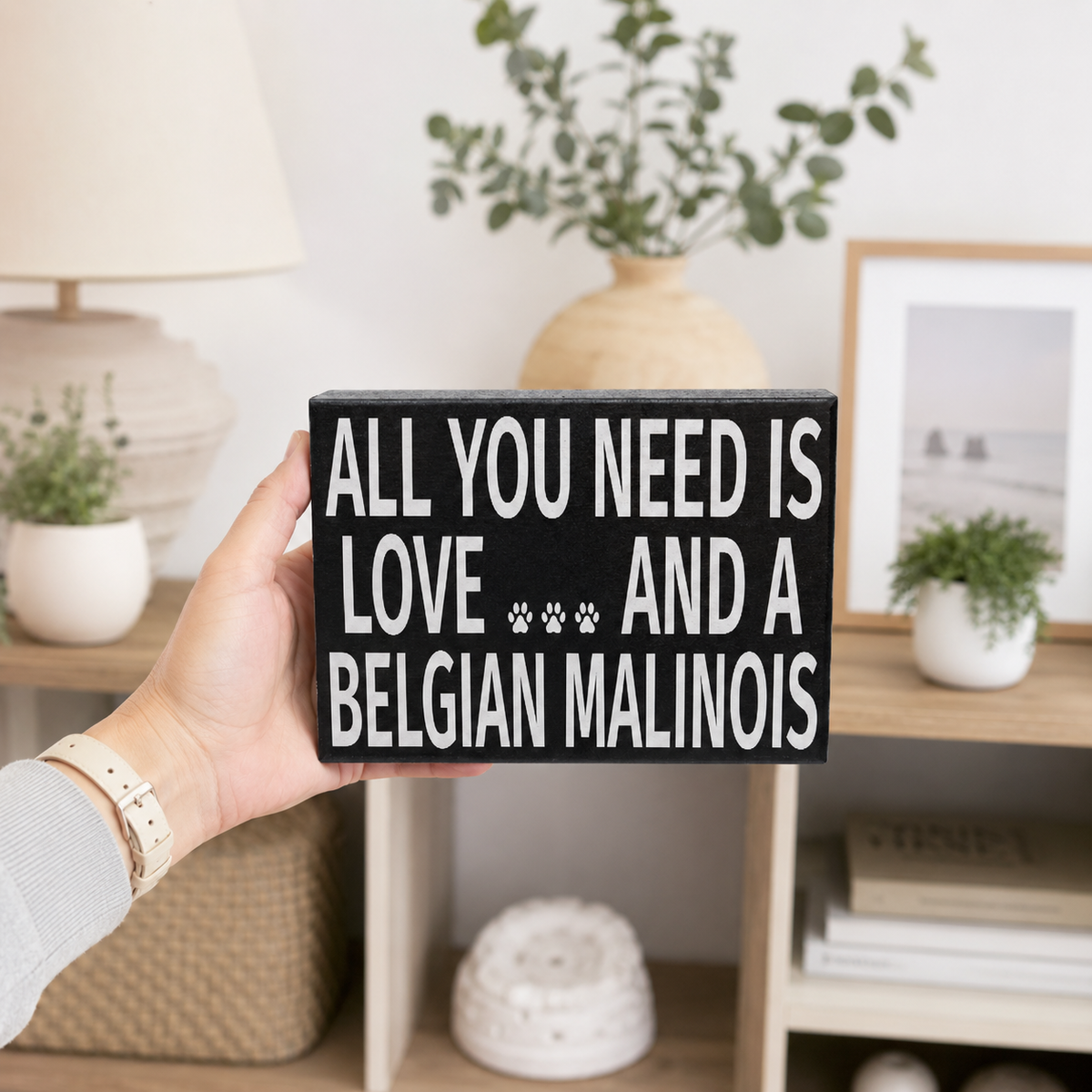 A hand holds a black rectangular wooden sign with white text that reads 'ALL YOU NEED IS LOVE .