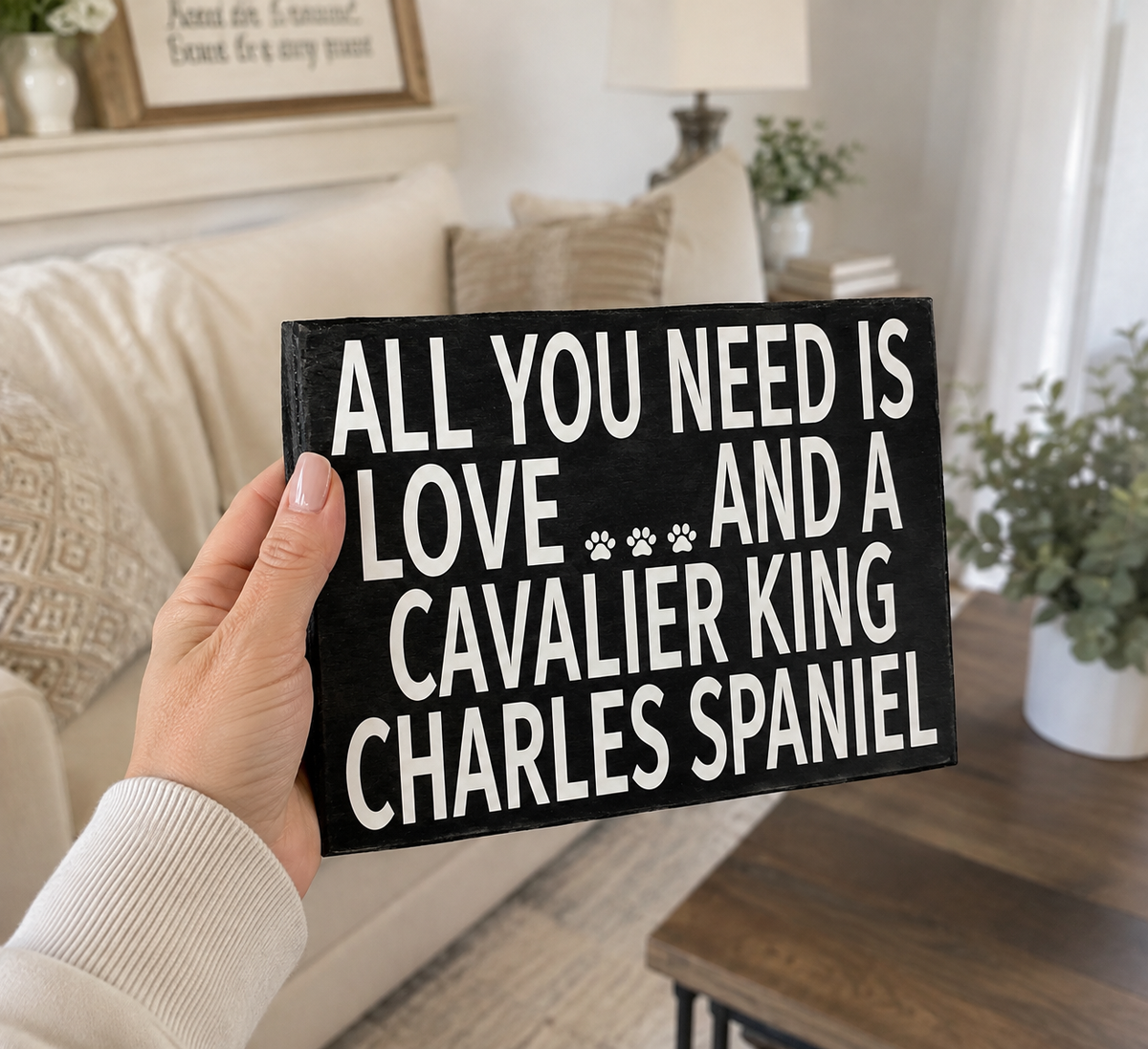 A hand holds a black rectangular sign with white text that reads, 'ALL YOU NEED IS LOVE AND A CAVALIER KING CHARLES SPANIEL' in a cozy living room setting.
