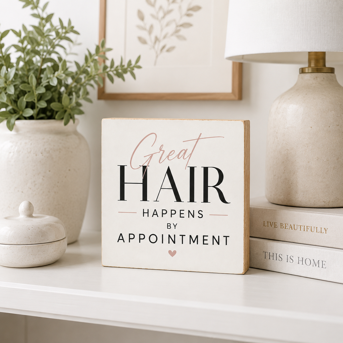 A square white decorative sign with the phrase 'Great HAIR HAPPENS BY APPOINTMENT' in black and blush pink lettering, styled on a white shelf beside a vase with greenery, a lamp, and stacked books.