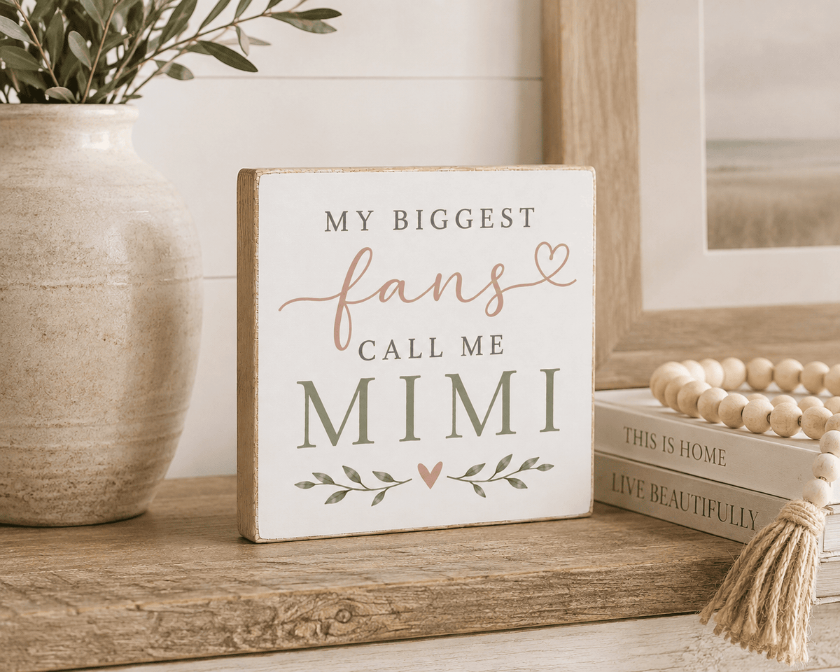 A square decorative sign with the phrase 'MY BIGGEST fans CALL ME MIMI' in dark gray and dusty rose lettering, styled on a rustic wooden surface next to a ceramic vase and books.