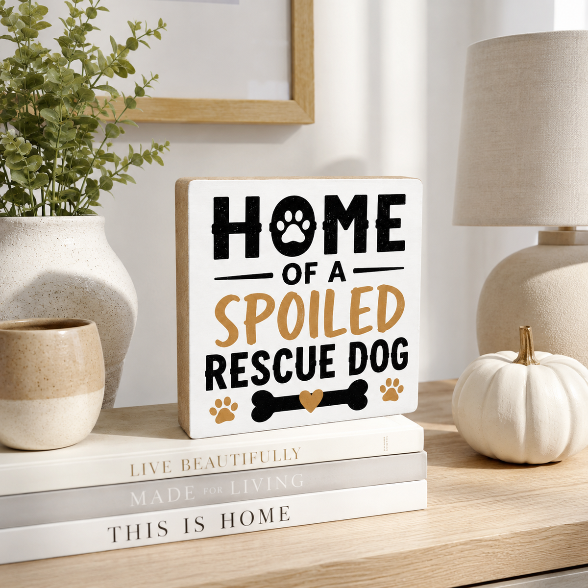 A square decorative sign reading “HOME OF A SPOILED RESCUE DOG” in black and tan lettering with paw and bone graphics, styled on a wooden table with books, a small pumpkin, a vase, and a lamp in a home interior setting.