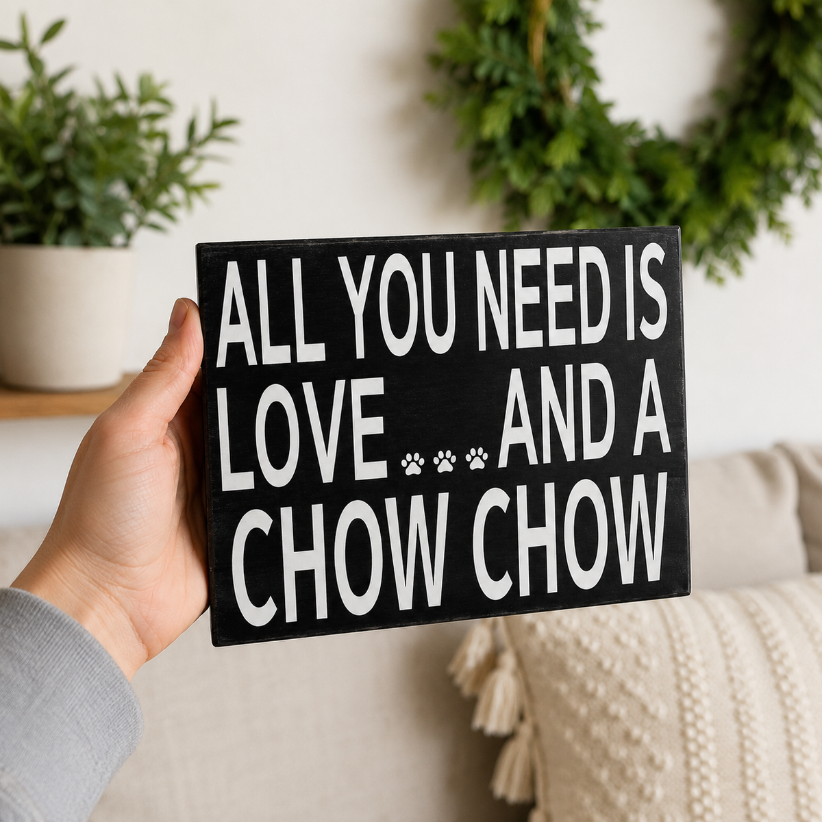 A hand holds a black rectangular wooden sign with white text that reads 'ALL YOU NEED IS LOVE.