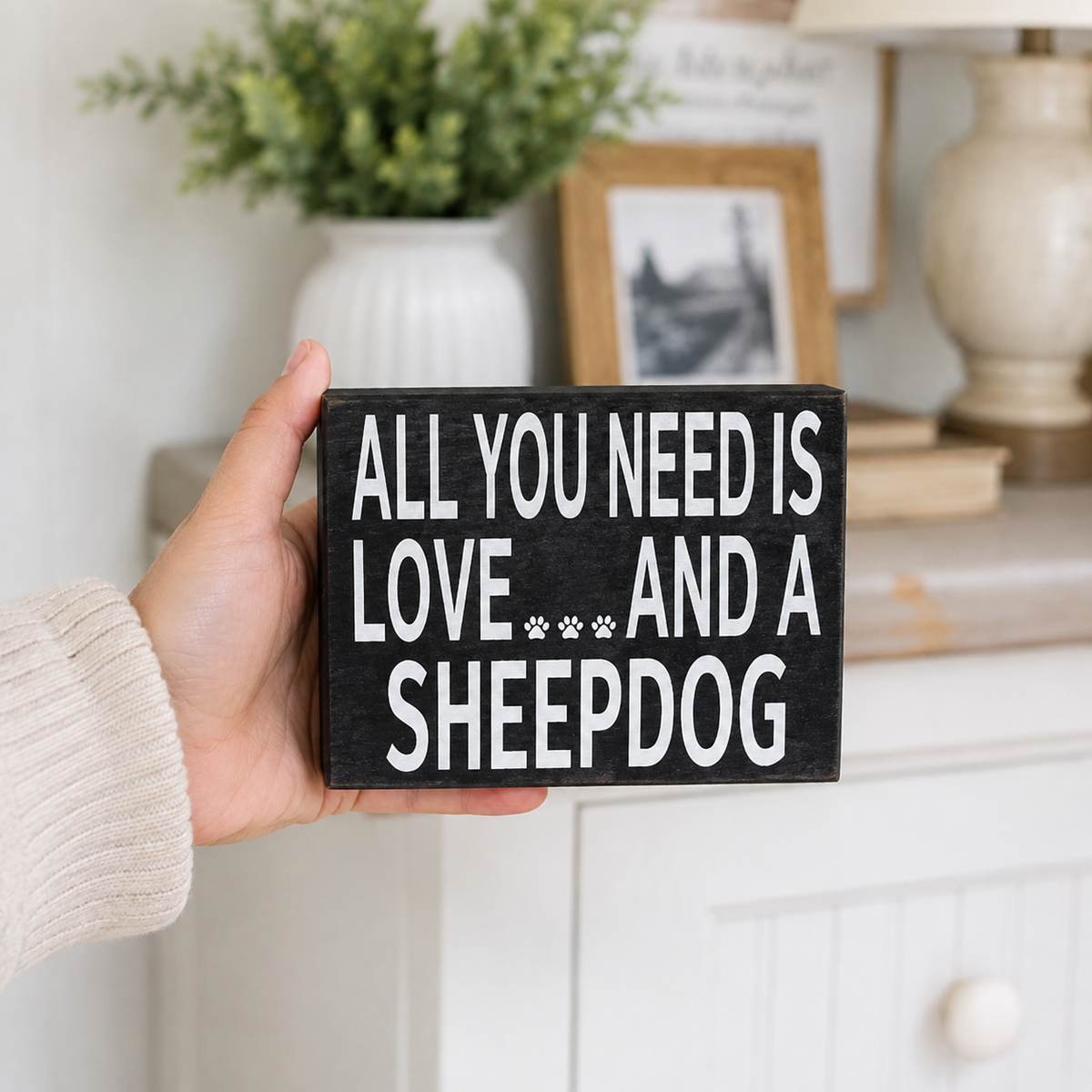 A hand holding a rectangular black wooden sign with white text that reads 'ALL YOU NEED IS LOVE *** AND A SHEEPDOG' against a softly blurred home interior background.