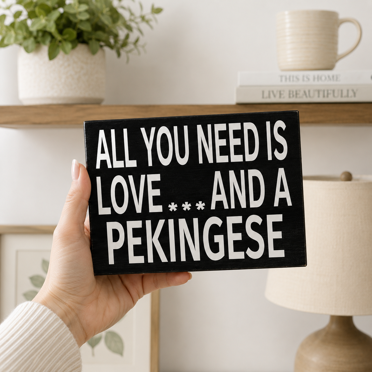 A hand holds a black rectangular sign with white text that reads 'ALL YOU NEED IS LOVE *** AND A PEKINGESE' in a cozy indoor setting with a shelf, mug, plant, and lamp in the background.