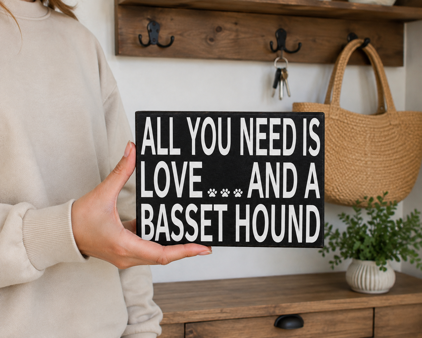 A person in a beige sweatshirt holds a black wooden sign with white text that reads 'ALL YOU NEED IS LOVE.