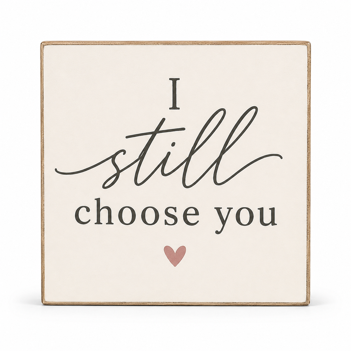 A square decorative sign with a cream background and a natural wood border. The sign features the phrase 'I still choose you' in black script and serif lettering, with a small dusty pink heart underneath.