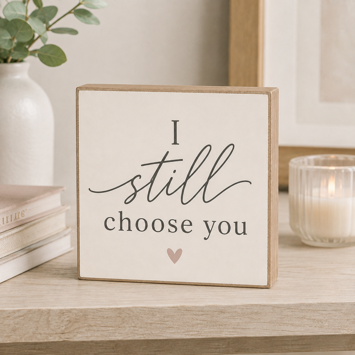A square wooden sign with a cream face and natural wood edges, printed with the phrase 'I still choose you' in black script and serif text, plus a small pink heart. The sign sits on a light wood table in a styled room setting with a white vase, books, and a lit candle in the background.