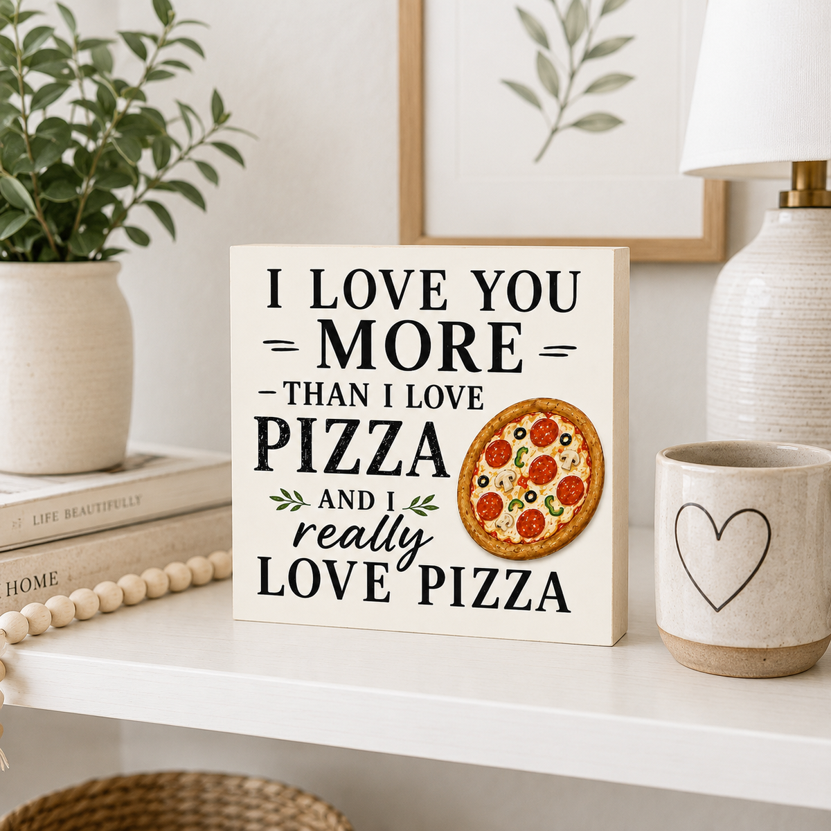 A decorative wooden sign on a white shelf reads “I LOVE YOU MORE THAN I LOVE PIZZA AND I really LOVE PIZZA” with a printed pizza illustration. The sign is styled in a home setting with a potted plant, books, a ceramic mug, and a lamp.