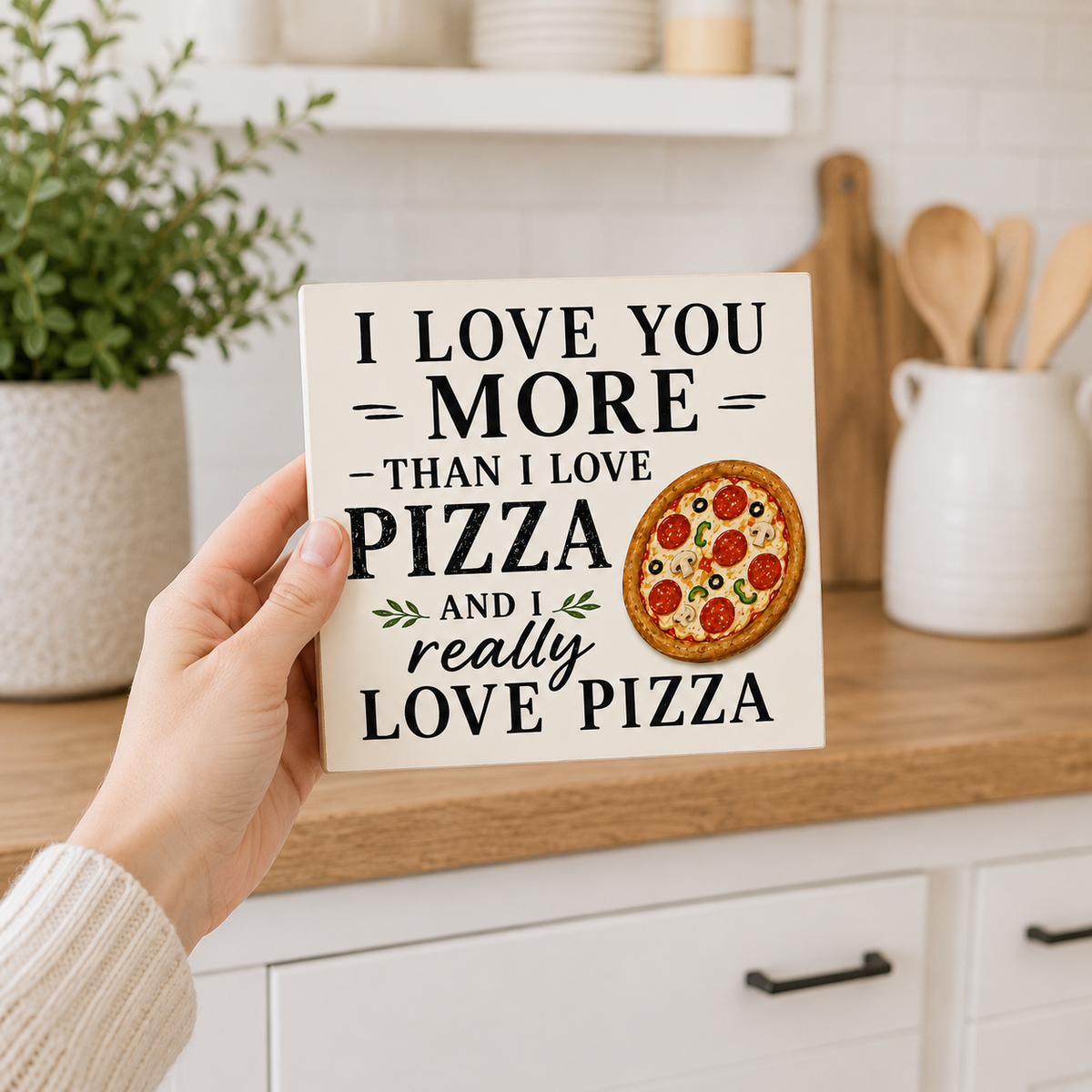 A hand holds a square ceramic sign with the text 'I LOVE YOU MORE THAN I LOVE PIZZA AND I really LOVE PIZZA' next to an illustration of a pepperoni pizza, set against a blurred kitchen background.