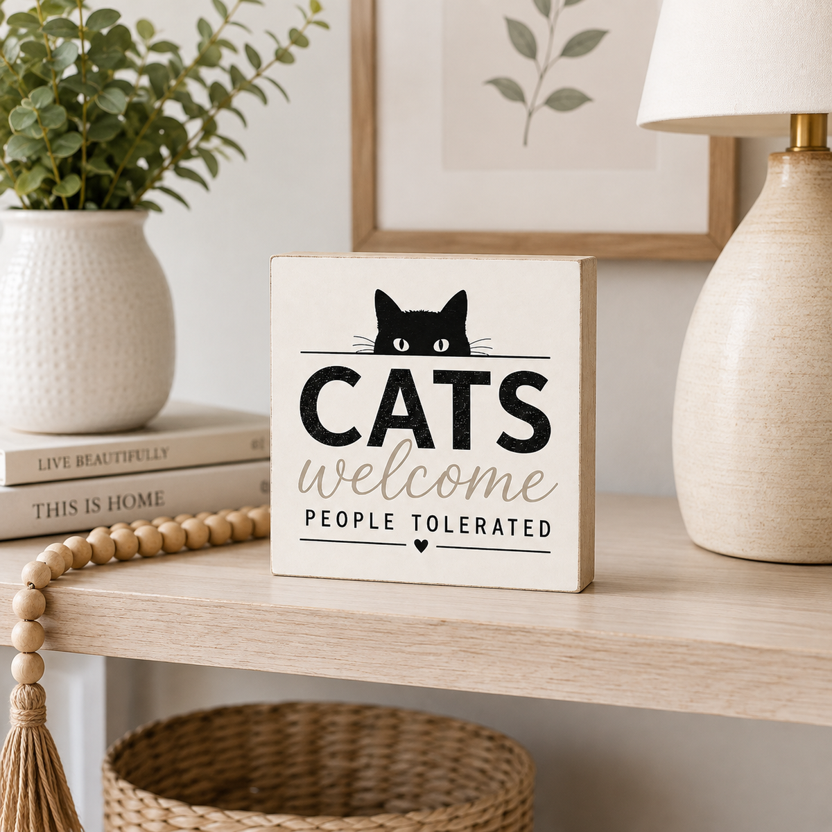 A square wooden sign with a black cat illustration and the text 'CATS welcome PEOPLE TOLERATED' sits on a light wood shelf in a styled home setting with a plant, lamp, and framed art.