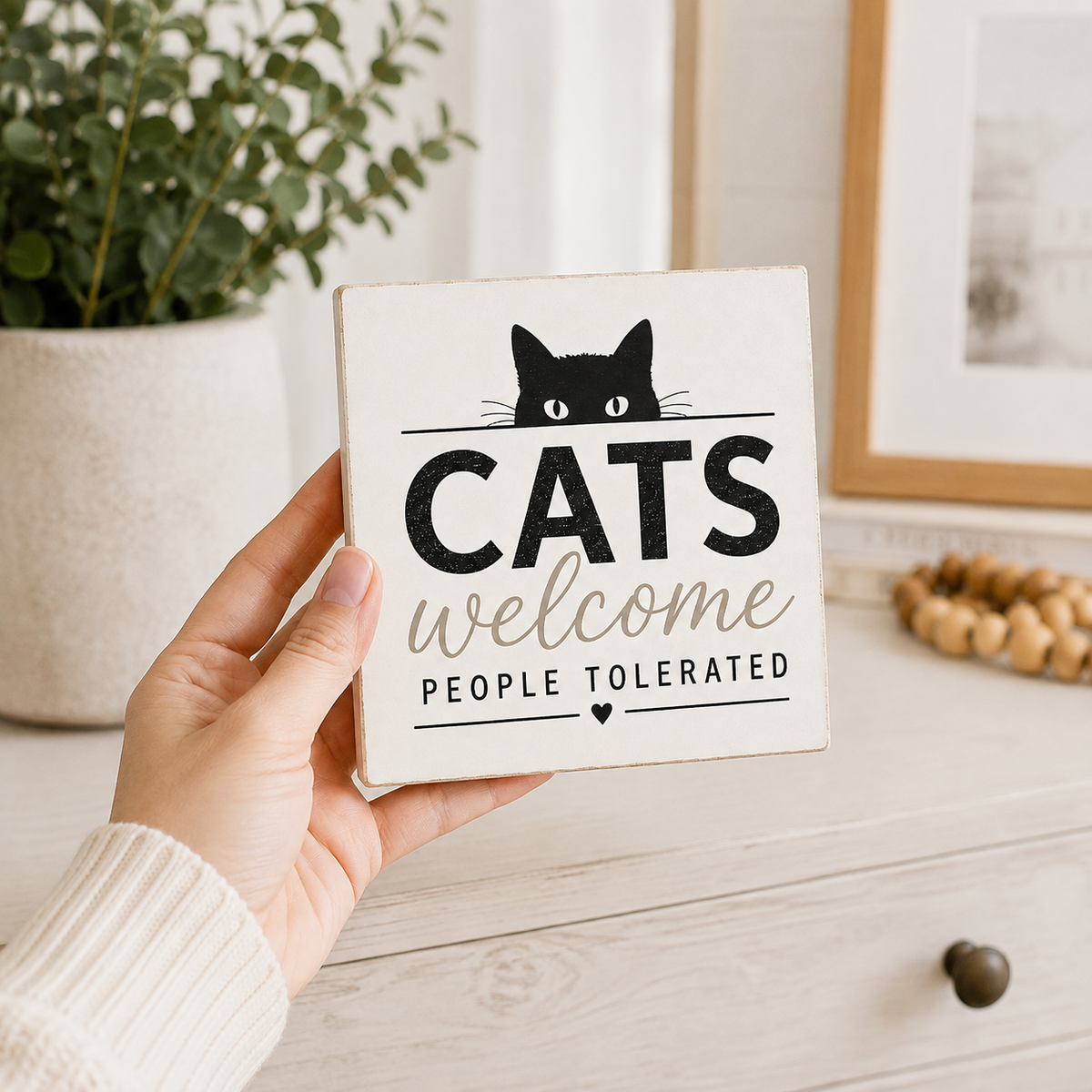 A hand holds a square wooden sign that reads 'CATS welcome PEOPLE TOLERATED' with a black cat peeking above the text. The sign is photographed in a bright, styled room setting with a potted plant, framed art, and wooden decor in the background.