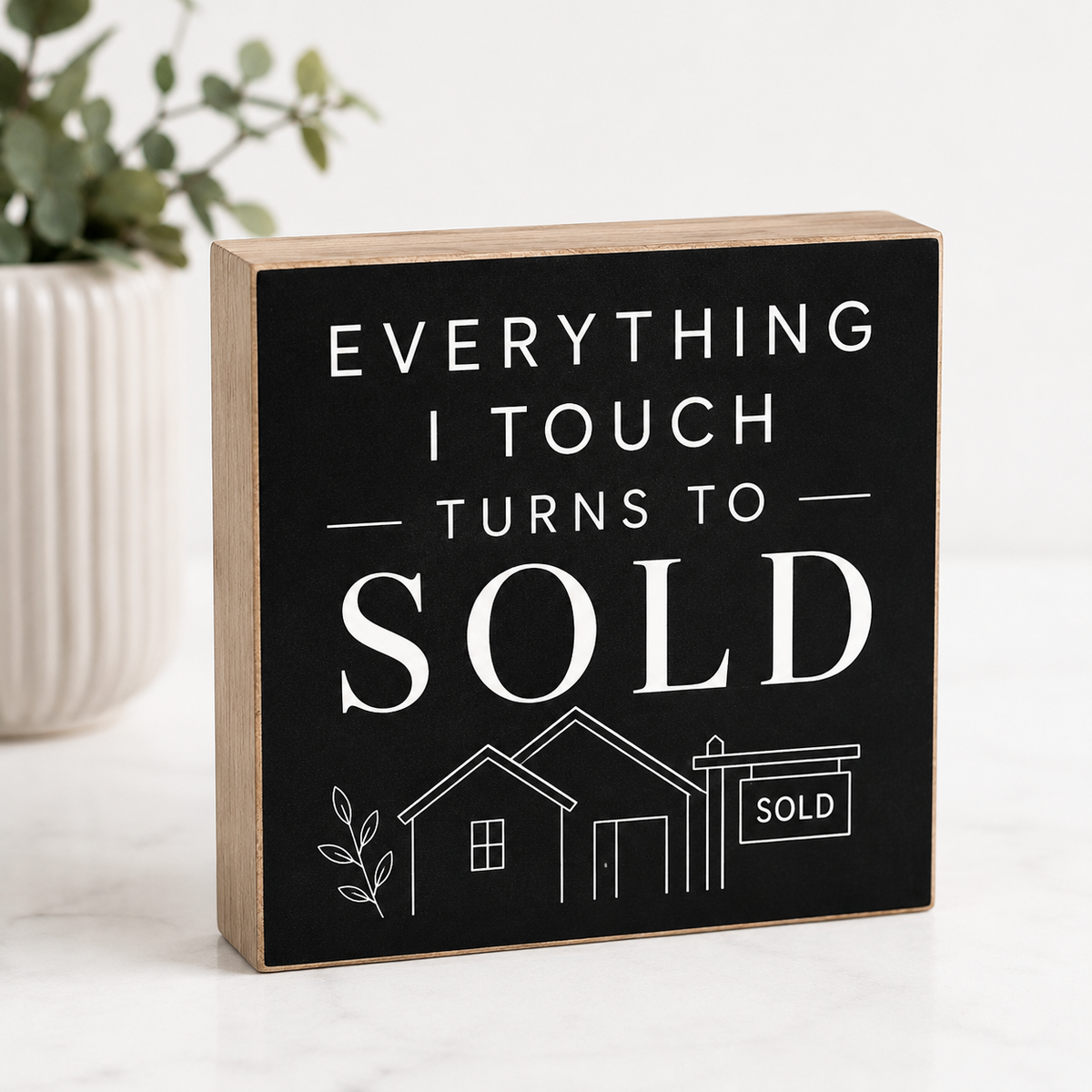 A rectangular wooden block sign with a black front panel and natural wood sides, printed with the phrase “EVERYTHING I TOUCH TURNS TO SOLD” in white text above a simple house and sold sign illustration. It is photographed on a light surface with a blurred plant in the background.