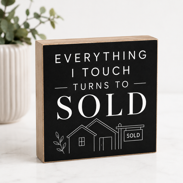 A rectangular wooden block sign with a black front panel and natural wood sides, printed with the phrase “EVERYTHING I TOUCH TURNS TO SOLD” in white text above a simple house and sold sign illustration. It is photographed on a light surface with a blurred plant in the background.