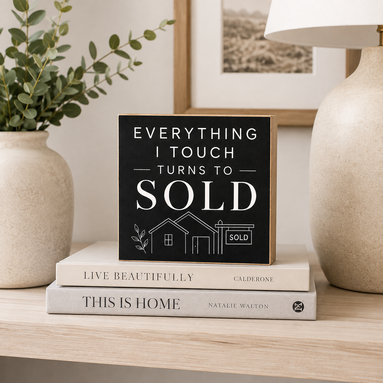 A square black decorative sign with white text reading 'EVERYTHING I TOUCH TURNS TO SOLD' and a small house-and-SOLD sign illustration, displayed on top of two books on a light wood shelf between a vase and a lamp.
