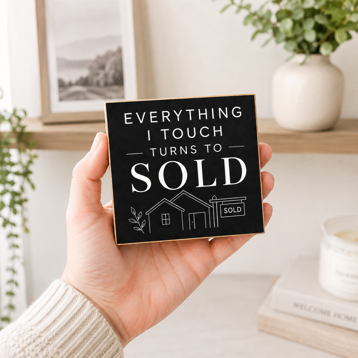 A hand holds a square black sign with gold trim that reads 'EVERYTHING I TOUCH TURNS TO SOLD' above a small house and garage illustration. The background shows a softly blurred home interior with a shelf, framed picture, plant, and candle.