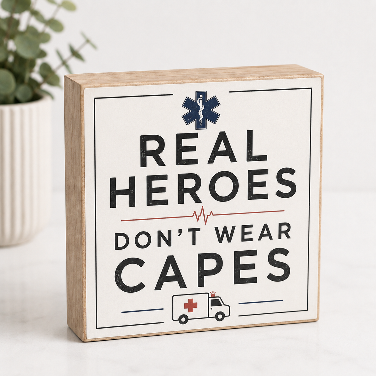 A square wooden sign with a light natural wood border and a white face, printed with the phrase 'REAL HEROES DON'T WEAR CAPES' in black text, plus a navy medical-style star and a small ambulance illustration. The sign is photographed on a light surface with a blurred plant in the background.