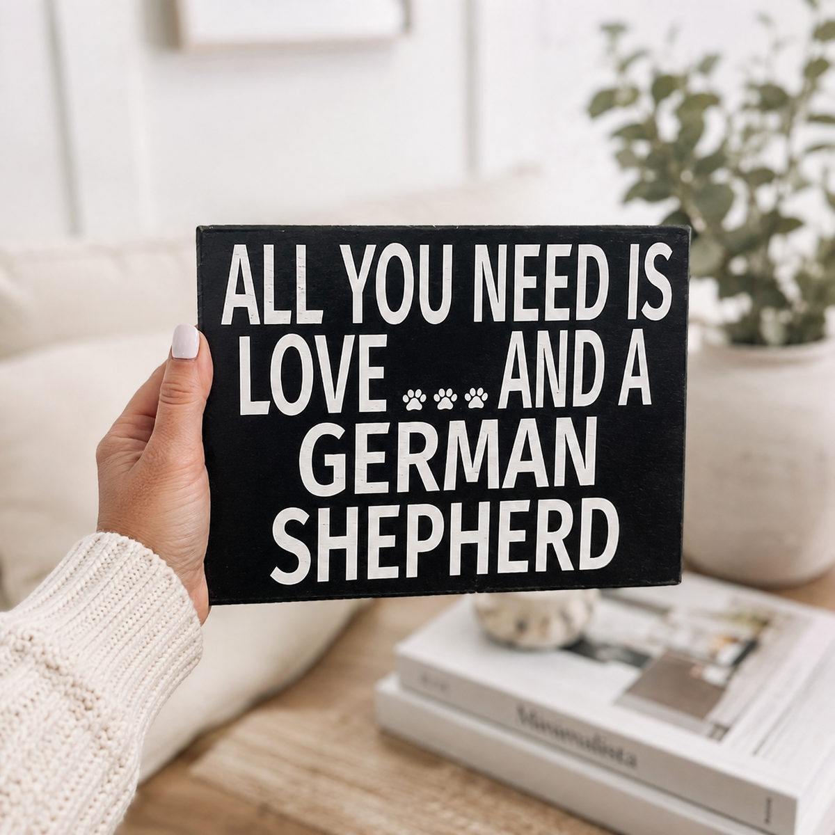 A hand holding a black square sign with white text that reads 'ALL YOU NEED IS LOVE *** AND A GERMAN SHEPHERD' in a cozy indoor setting with a plant and books in the background.
