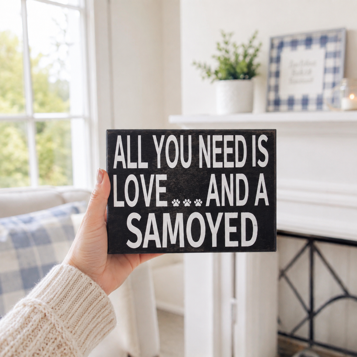 A hand wearing a cream knit sweater holds a black rectangular wooden sign with white text that reads 'ALL YOU NEED IS LOVE.