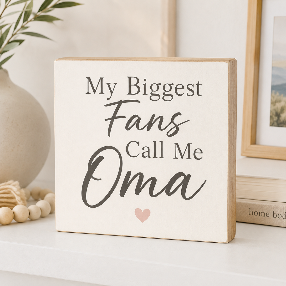 A square wooden sign with a cream face and natural wood edges, printed with the phrase 'My Biggest Fans Call Me Oma' in dark gray lettering and a small pink heart. The sign is styled on a white shelf next to a ceramic vase, wooden beads, and a framed picture.