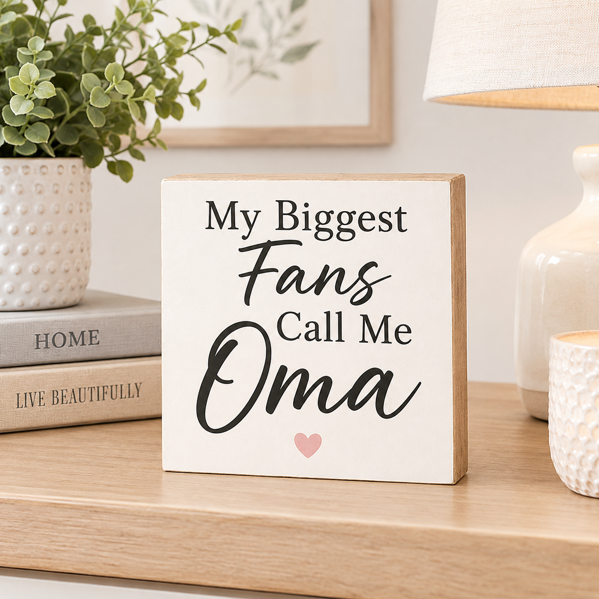 A square wooden sign with the phrase 'My Biggest Fans Call Me Oma' in black lettering, accented with a small pink heart, displayed on a wooden shelf in a styled home setting.