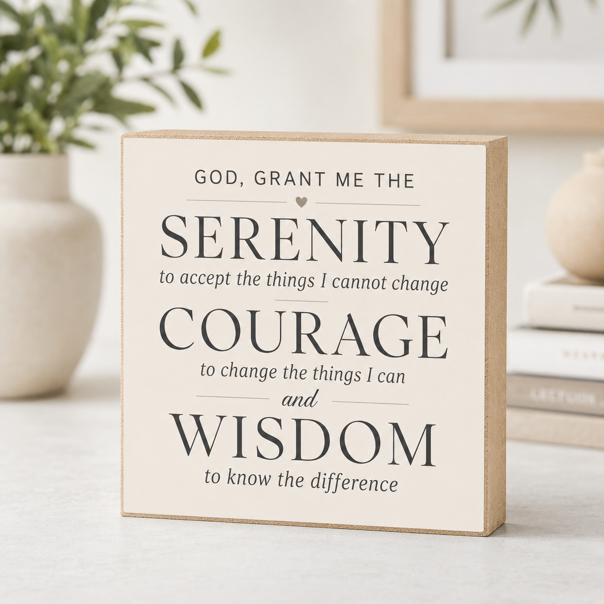 A square decorative wooden sign with a cream face and natural wood edges, printed with the Serenity Prayer in black serif and script text. The sign is photographed on a light tabletop with a softly blurred home interior background including a plant, books, and framed art.