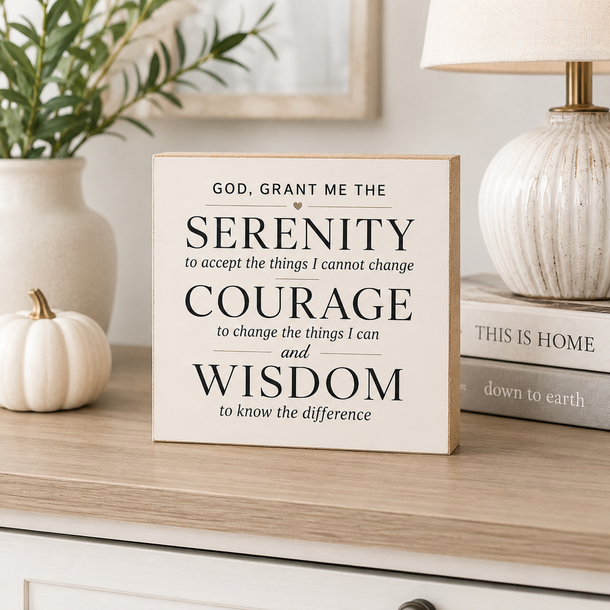 A square wooden sign with black text reading a Serenity prayer, styled on a light wood dresser next to a white ceramic vase with greenery, a small white pumpkin, a lamp, and stacked books.