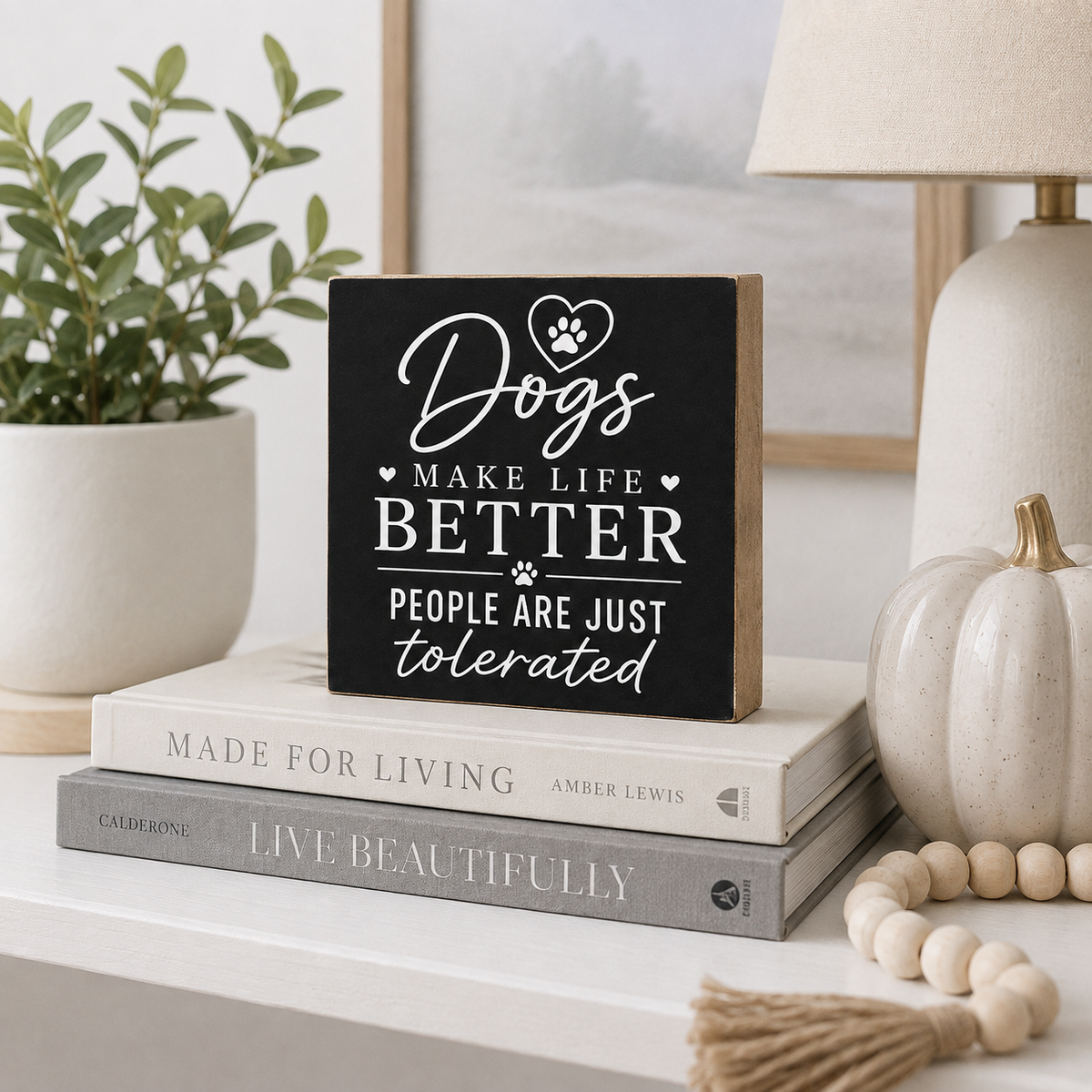 A square black wooden sign with white text reading 'Dogs MAKE LIFE BETTER PEOPLE ARE JUST tolerated' styled on a shelf with books, a plant, a lamp, and a small pumpkin.