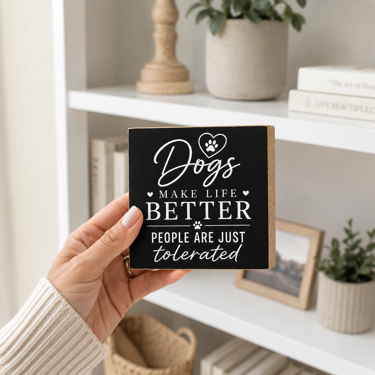 A hand holding a square black decorative wooden sign with white text that reads 'Dogs MAKE LIFE BETTER PEOPLE ARE JUST tolerated' in a cozy indoor setting with a white bookshelf in the background.