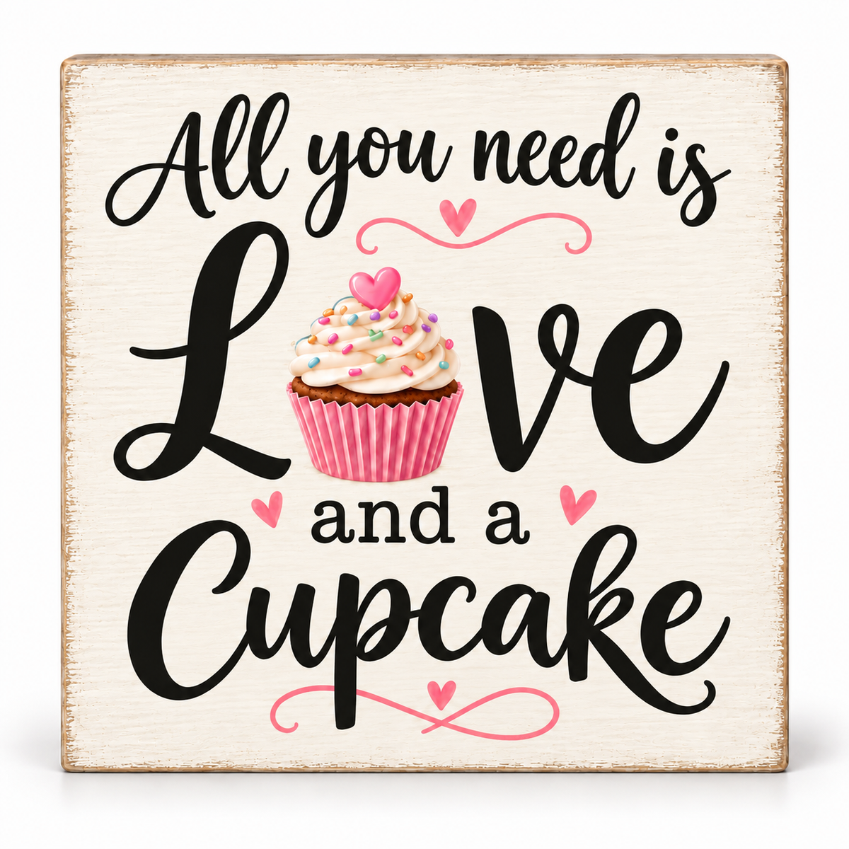 A square decorative sign with a distressed cream background and rustic brown edges. The sign features the phrase 'All you need is Love and a Cupcake' in black script and serif lettering, with a pink cupcake illustration in the center and small pink hearts and swirl accents.
