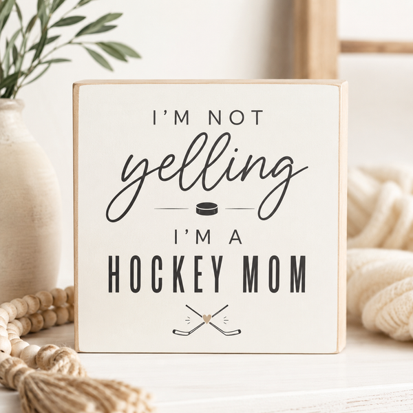 A square decorative sign with a light wood border and cream face reading “I’M NOT yelling I’M A HOCKEY MOM” in black lettering, with a small hockey puck and crossed hockey sticks graphic. The sign is styled on a tabletop with a vase, rope, and soft neutral background.