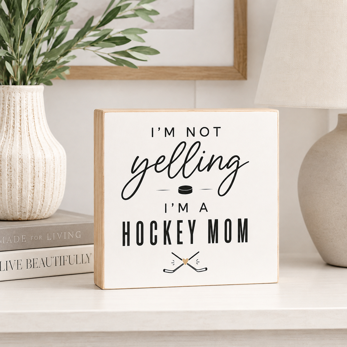 A square white decorative sign with a light wood edge and black text reading “I'M NOT yelling I'M A HOCKEY MOM,” with a small hockey puck and crossed hockey sticks graphic. The sign is styled on a tabletop in a home setting next to a vase with greenery and a lamp.