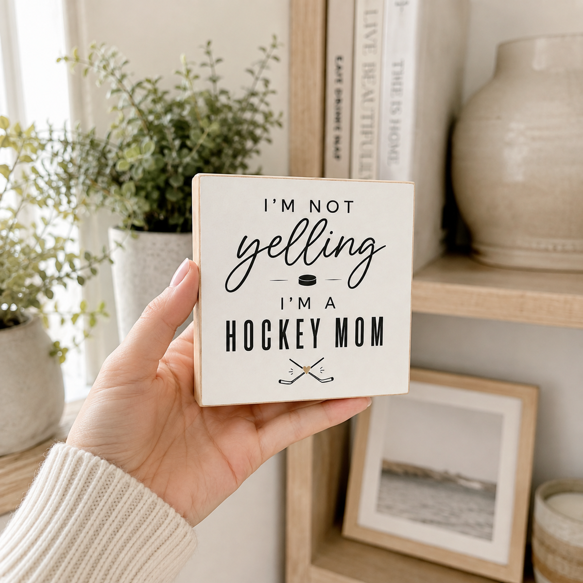 A hand holding a square white decorative sign that reads 'I'M NOT yelling I'M A HOCKEY MOM' with crossed hockey sticks underneath, set against a cozy indoor background with plants and shelves.