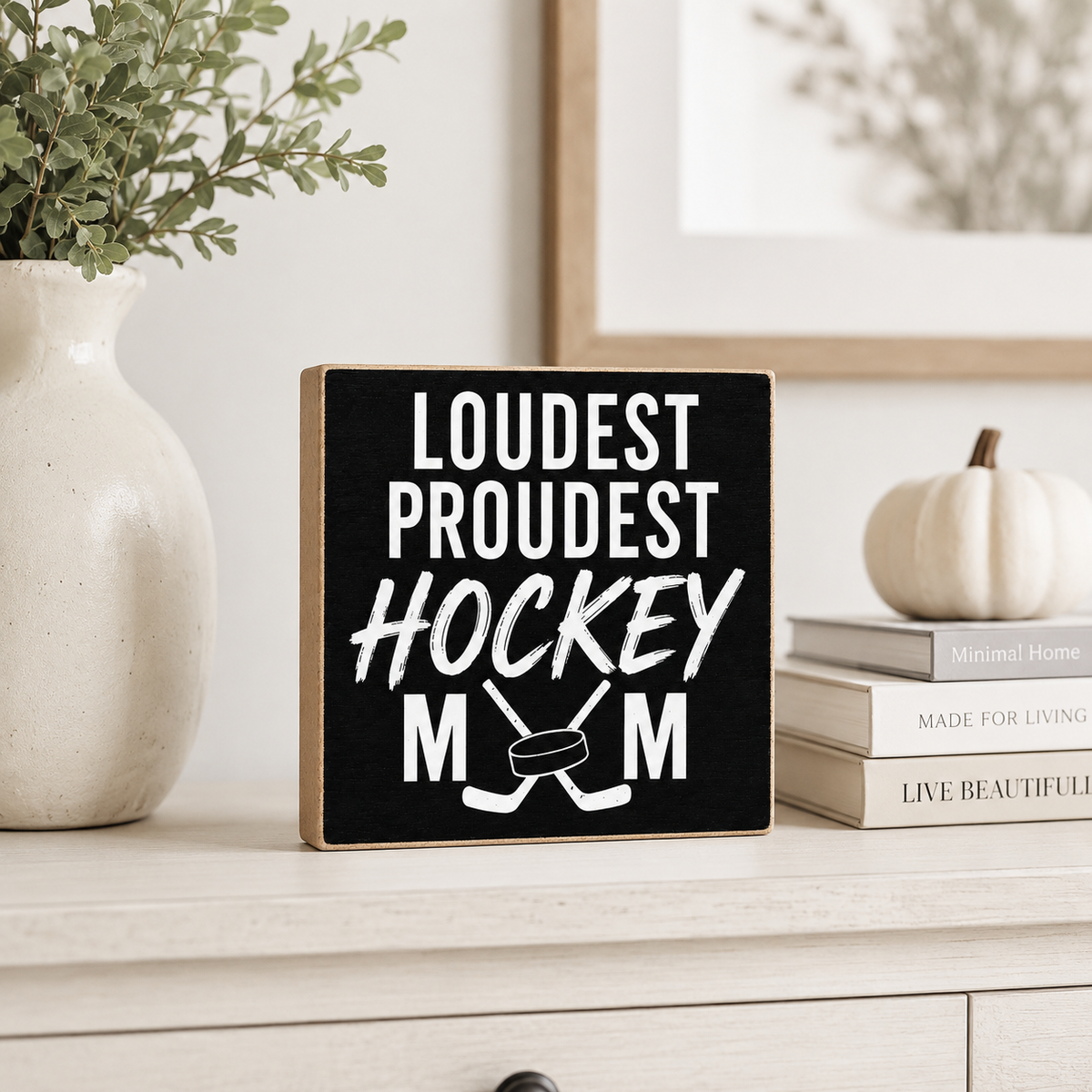 A square black wooden sign with white text reading 'LOUST PROUDEST HOCKEY MUM' and crossed hockey sticks, displayed on a light wood dresser in a styled room setting with a vase, books, and a small pumpkin.