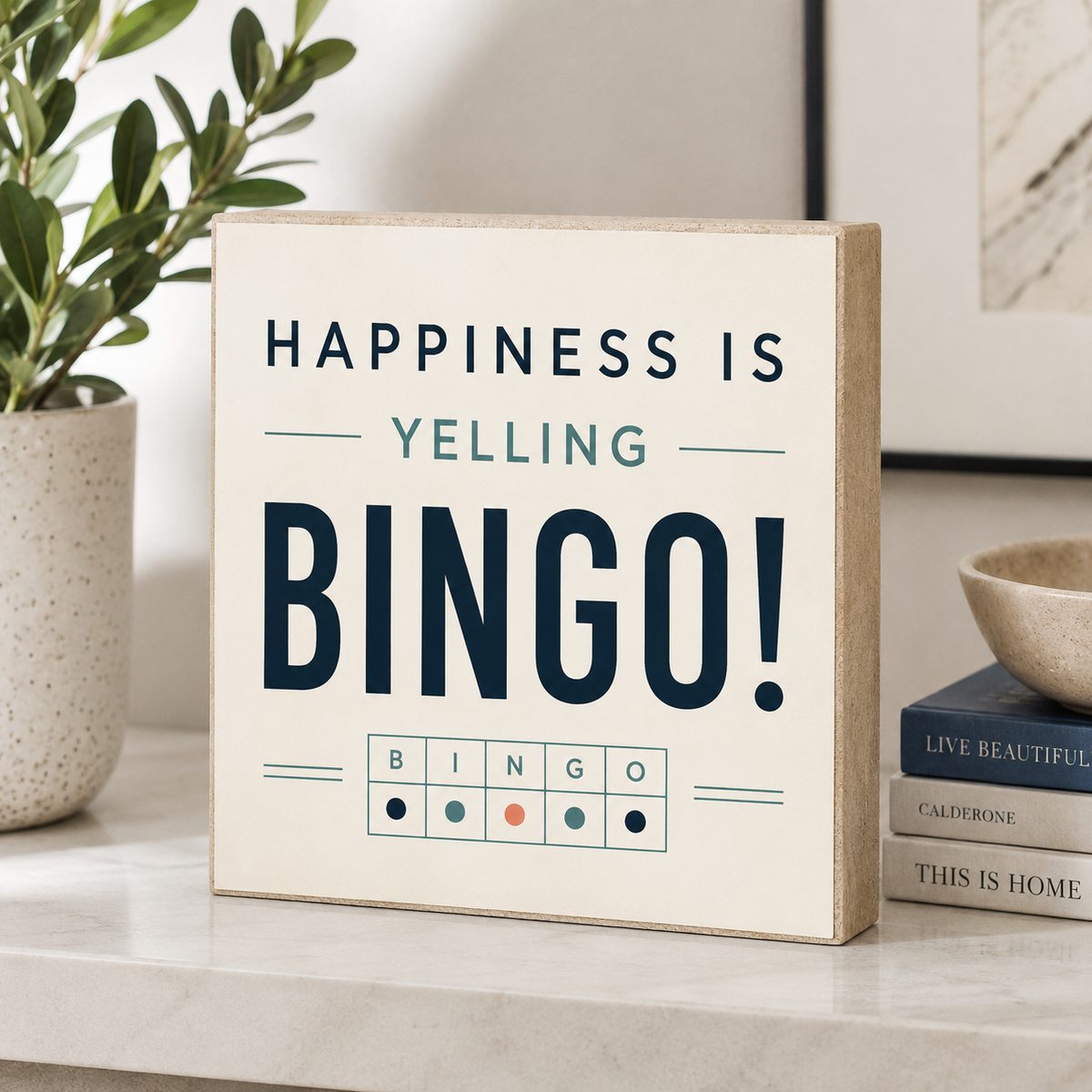 A square decorative sign with the phrase “HAPPINESS IS YELLING BINGO!” in dark navy and teal lettering on a warm cream background, styled on a light marble surface with a potted plant and books nearby.
