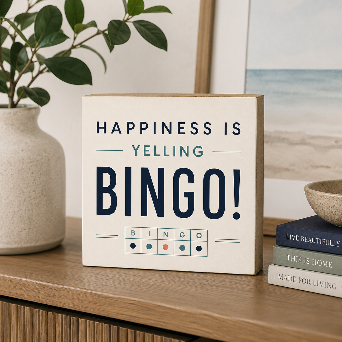 A square decorative sign with a light wood border and an off-white face reading “HAPPINESS IS YELLING BINGO!” in dark navy and teal lettering, styled on a wooden shelf beside a vase with green leaves and stacked books.