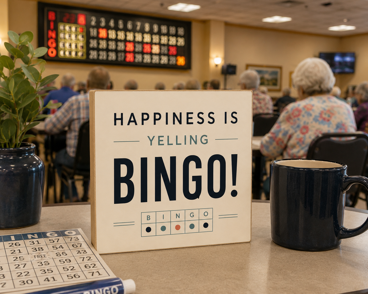A square decorative sign reading 'HAPPINESS IS YELLING BINGO!' sits on a table in a bingo lounge, with a bingo card, a dark mug, and a potted plant in the foreground and people seated in the background.