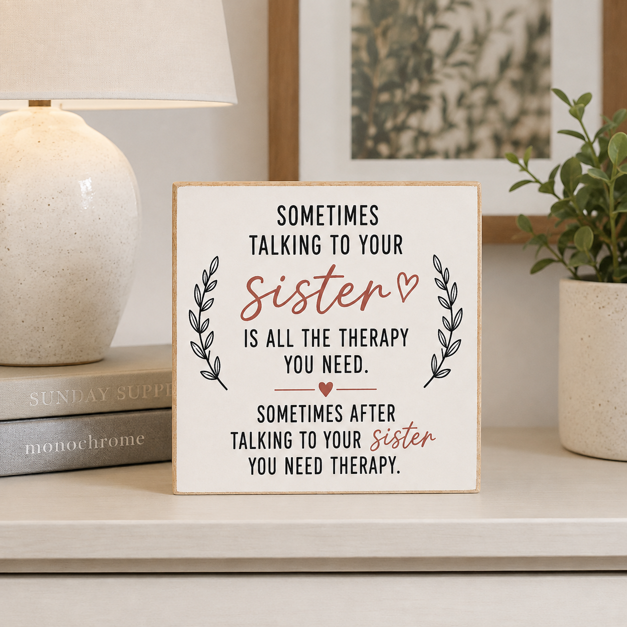 A square decorative sign with printed therapy-themed text for a sister, styled on a tabletop beside a lamp, books, and a potted plant.