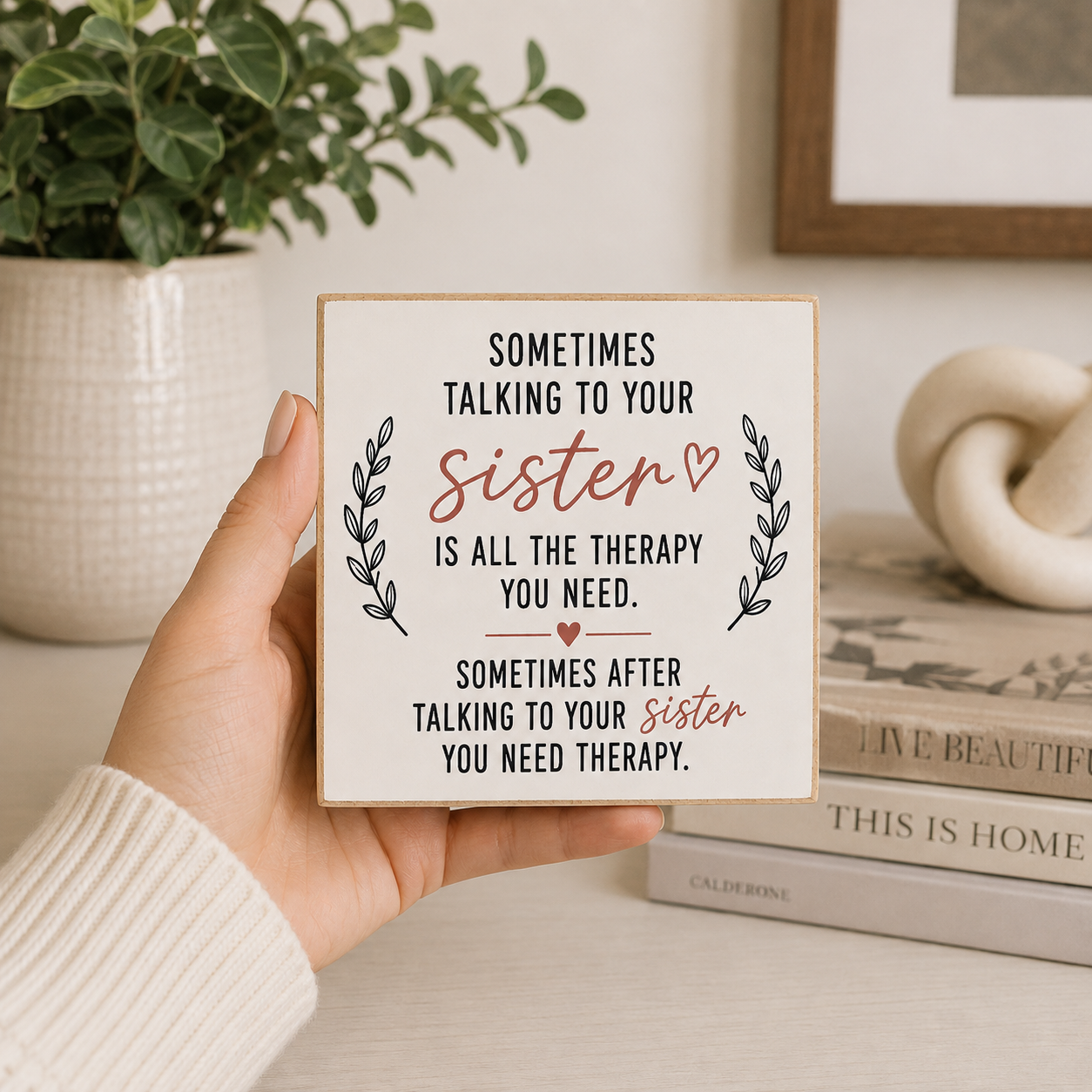 A hand holds a square wooden sign with a humorous quote about talking to a sister. The sign has a cream background with black text, black leaf illustrations, and the word 'sister' highlighted in a dusty rose color.