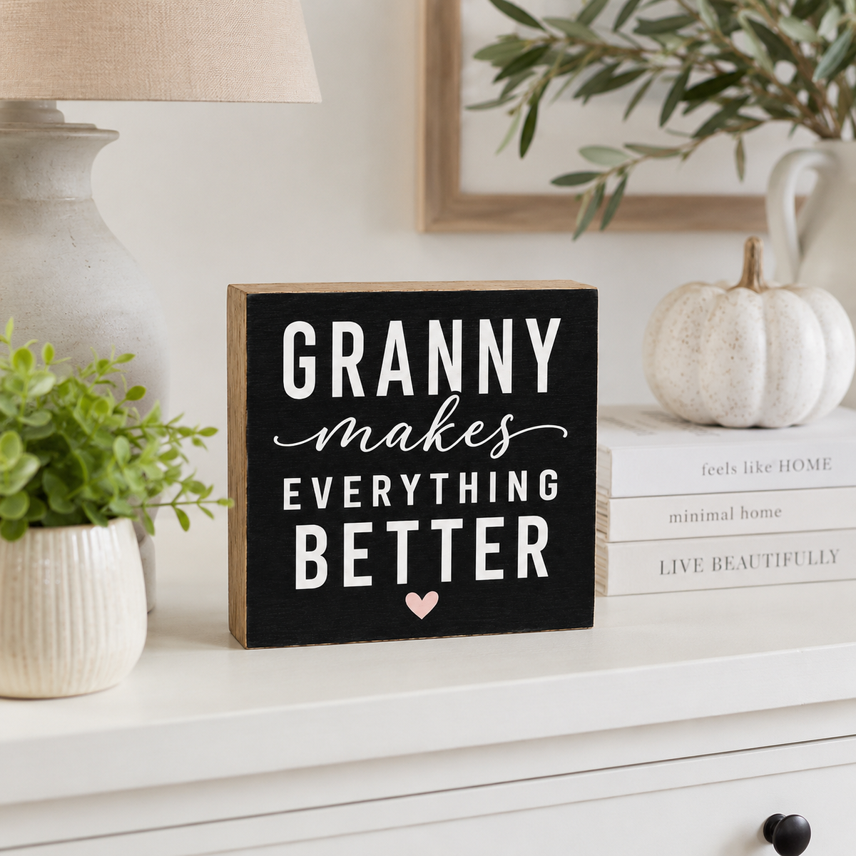 A rectangular black wooden sign with natural wood edges displays the phrase 'GRANNY makes EVERYTHING BETTER' in white lettering, accented by a small pink heart at the bottom. The sign is styled on a white dresser alongside a lamp, books, a small white pumpkin, and potted plants.