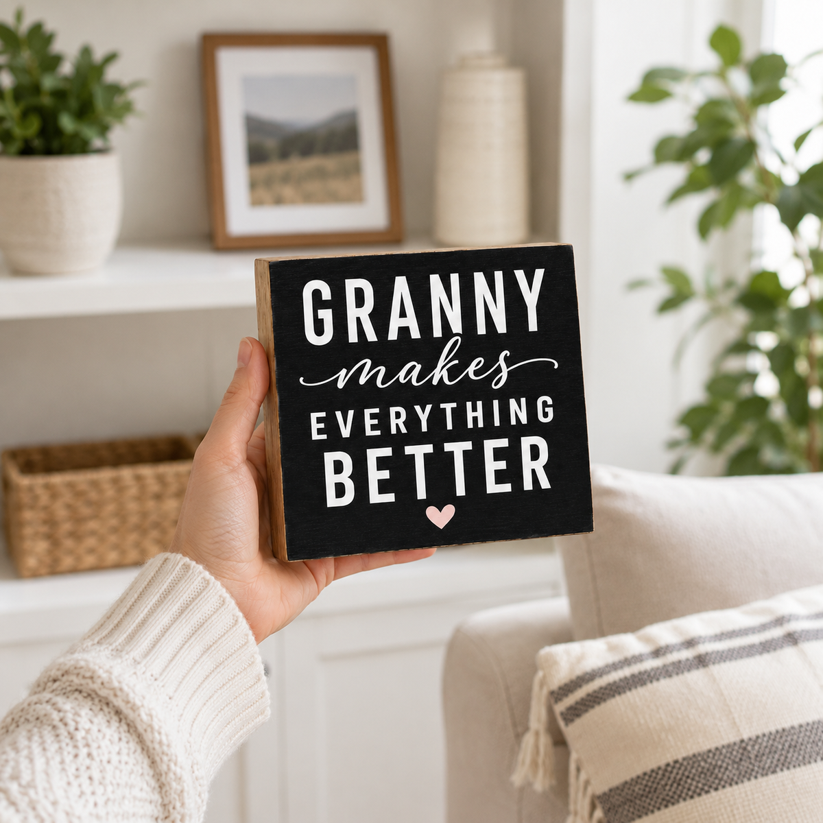 A hand in a cream knit sweater holds a square wooden sign with a black face and white text that reads “GRANNY makes EVERYTHING BETTER” with a small pink heart. The background shows a cozy living room setting with a sofa, plants, and shelves.