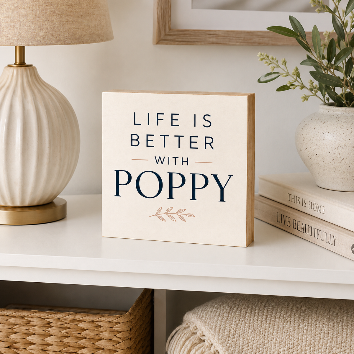 A square cream-colored wooden sign with the text 'LIFE IS BETTER WITH POPPY' in navy blue, styled on a white shelf next to a lamp, books, and a vase.