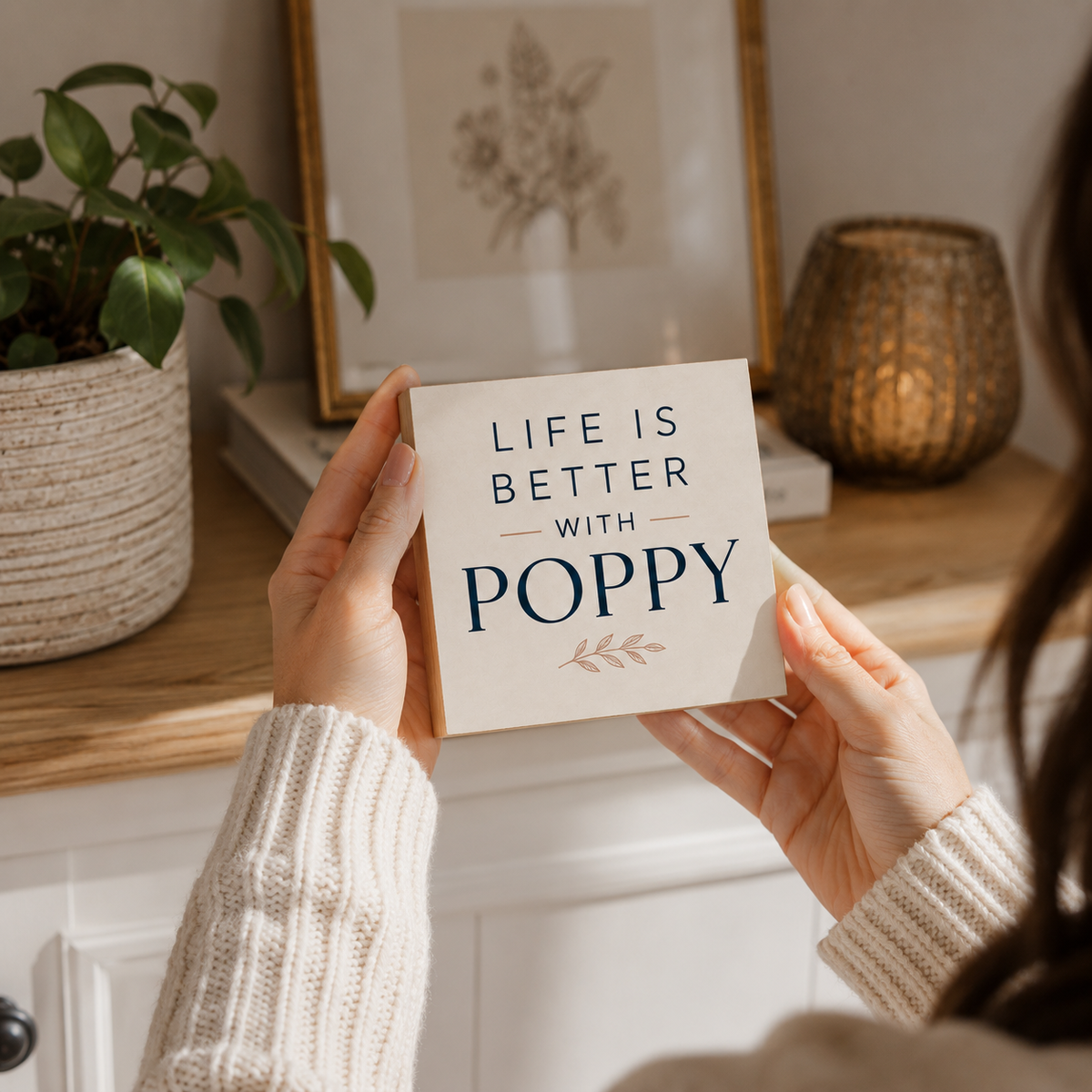 A person holding a square decorative sign that reads 'LIFE IS BETTER WITH POPPY' in navy text on a cream background, with a small leaf motif below. The photo is taken in a warm, styled interior with a plant, framed art, and a woven candle holder in the background.