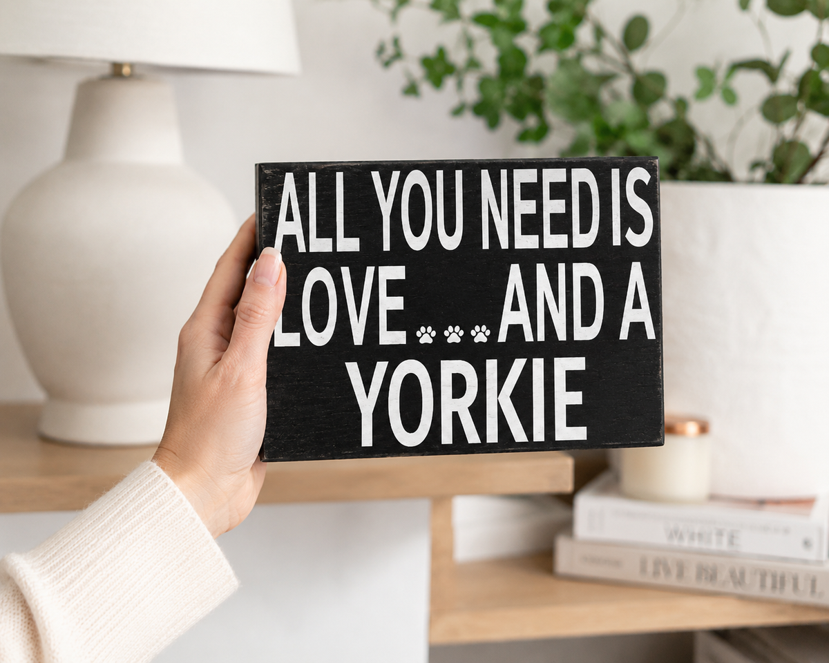 A hand holds a black rectangular wooden sign with white text reading 'ALL YOU NEED IS LOVE AND A YORKIE' with small paw prints. The background shows a softly blurred home interior with a white lamp, green plant, and books on a shelf.