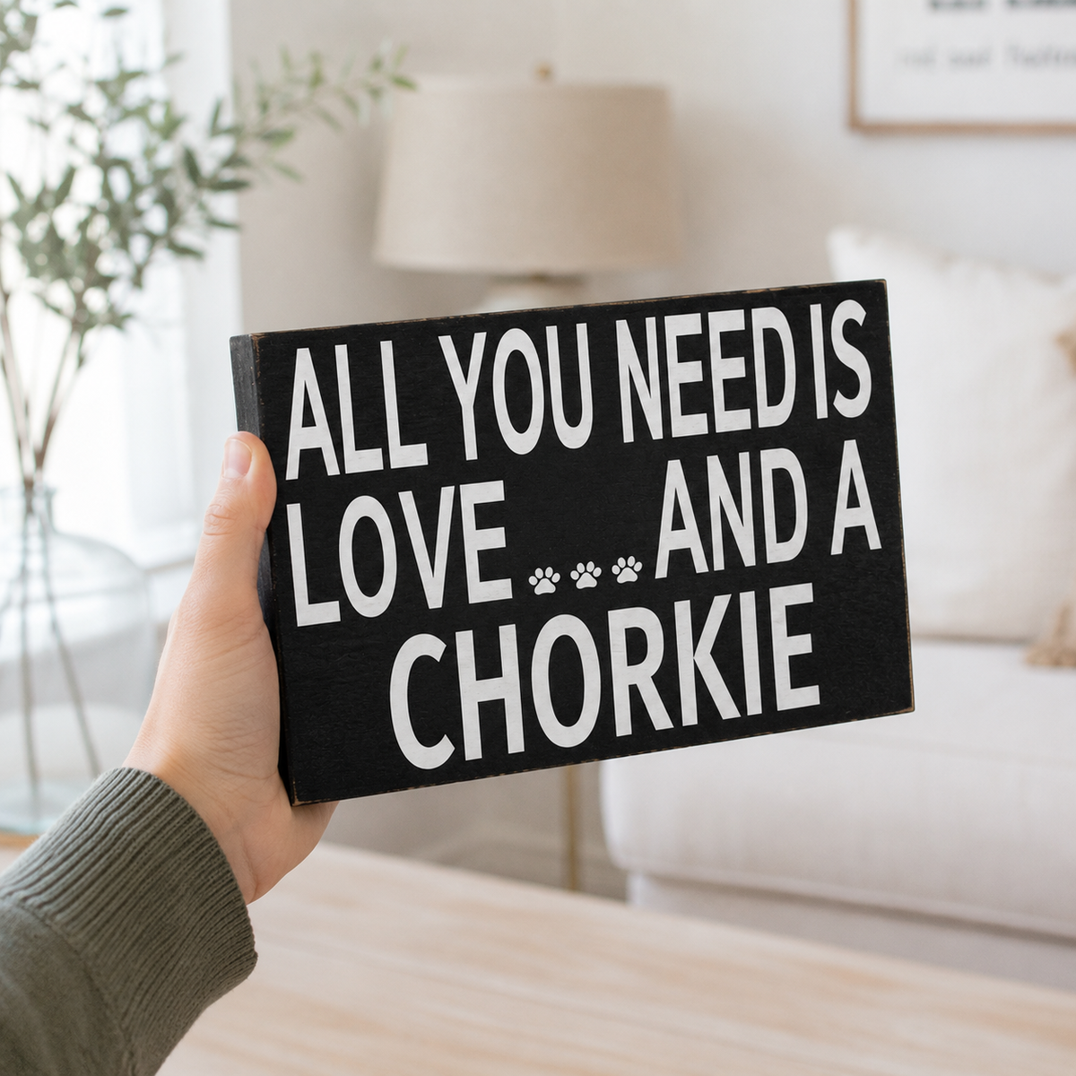 A hand holds a black rectangular wooden sign with bold white text reading 'ALL YOU NEED IS LOVE.