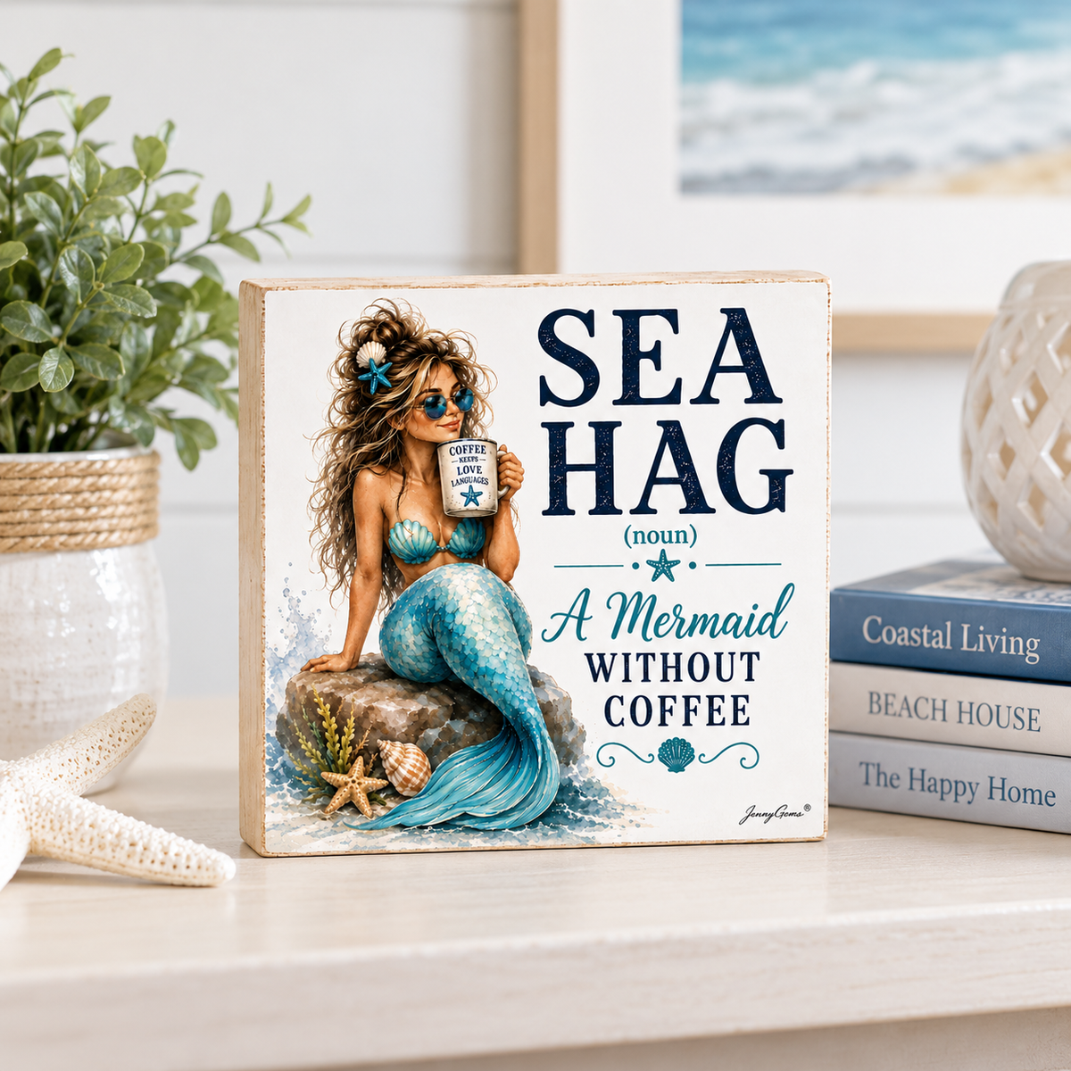A square decorative sign with a mermaid illustration and the text 'SEA HAG (noun) A Mermaid WITHOUT COFFEE' displayed on a light tabletop with a plant, books, and framed art in the background.
