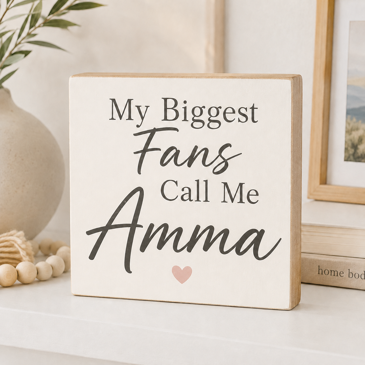 A square wooden sign with a cream face and natural wood edges, printed with the phrase 'My Biggest Fans Call Me Amma' in dark gray lettering and a small pink heart. The sign is styled on a white shelf with a ceramic vase, wooden beads, and a picture frame in the background.