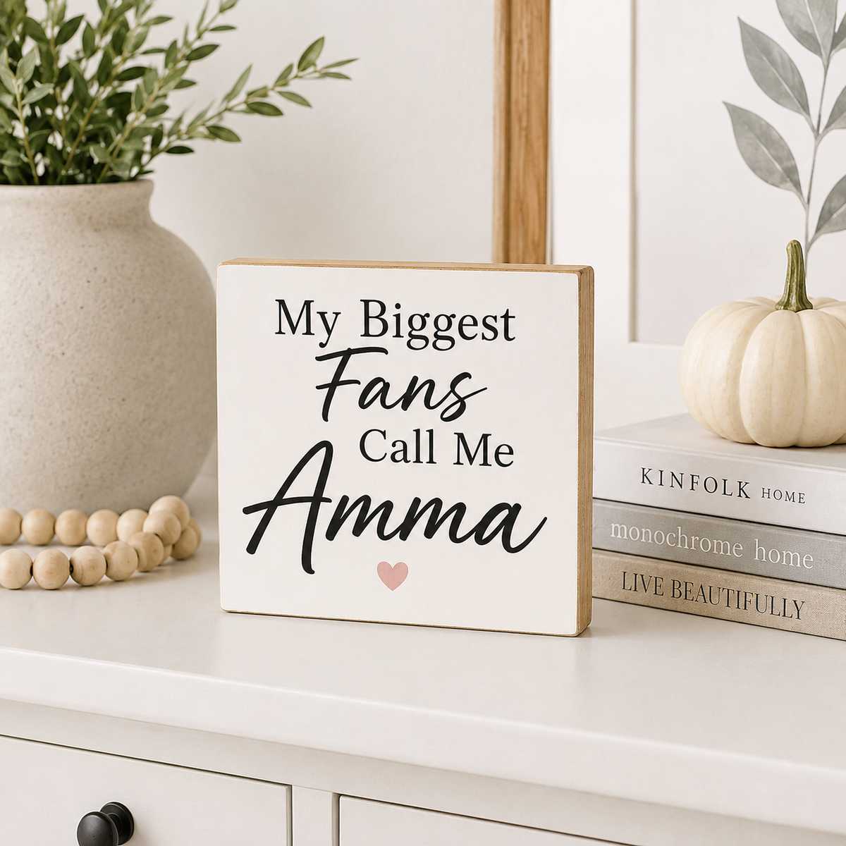 A square wooden sign with a white face and natural wood edges reads 'My Biggest Fans Call Me Amma' in black script and serif lettering, with a small pink heart at the bottom. It is styled on a white cabinet in a home setting next to a ceramic vase with greenery, wooden beads, a small white pumpkin, and a stack of books.