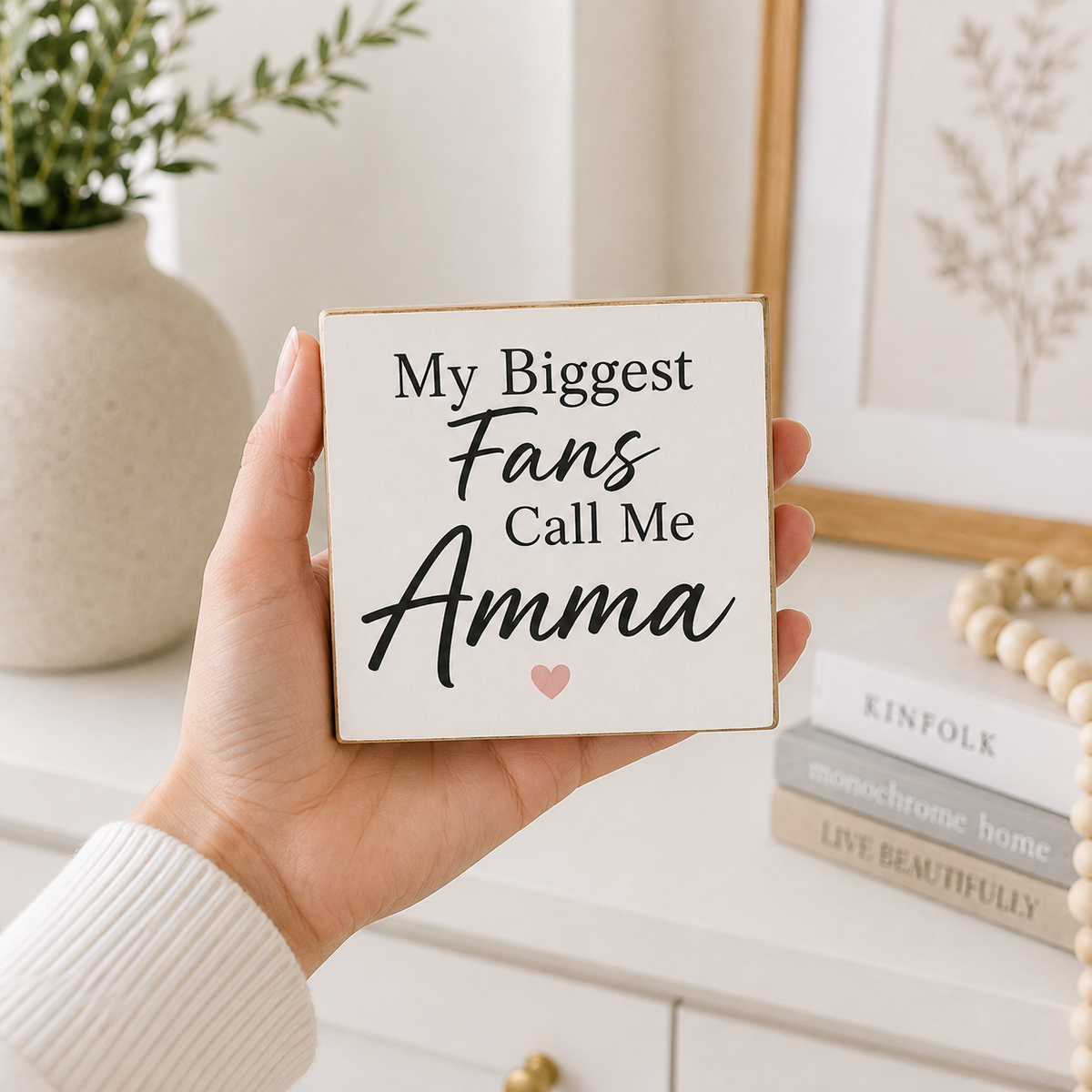 A hand holds a square white wooden sign with the text 'My Biggest Fans Call Me Amma' in black script and serif lettering, with a small pink heart at the bottom. The background shows a softly styled home interior with a vase, framed art, and books.