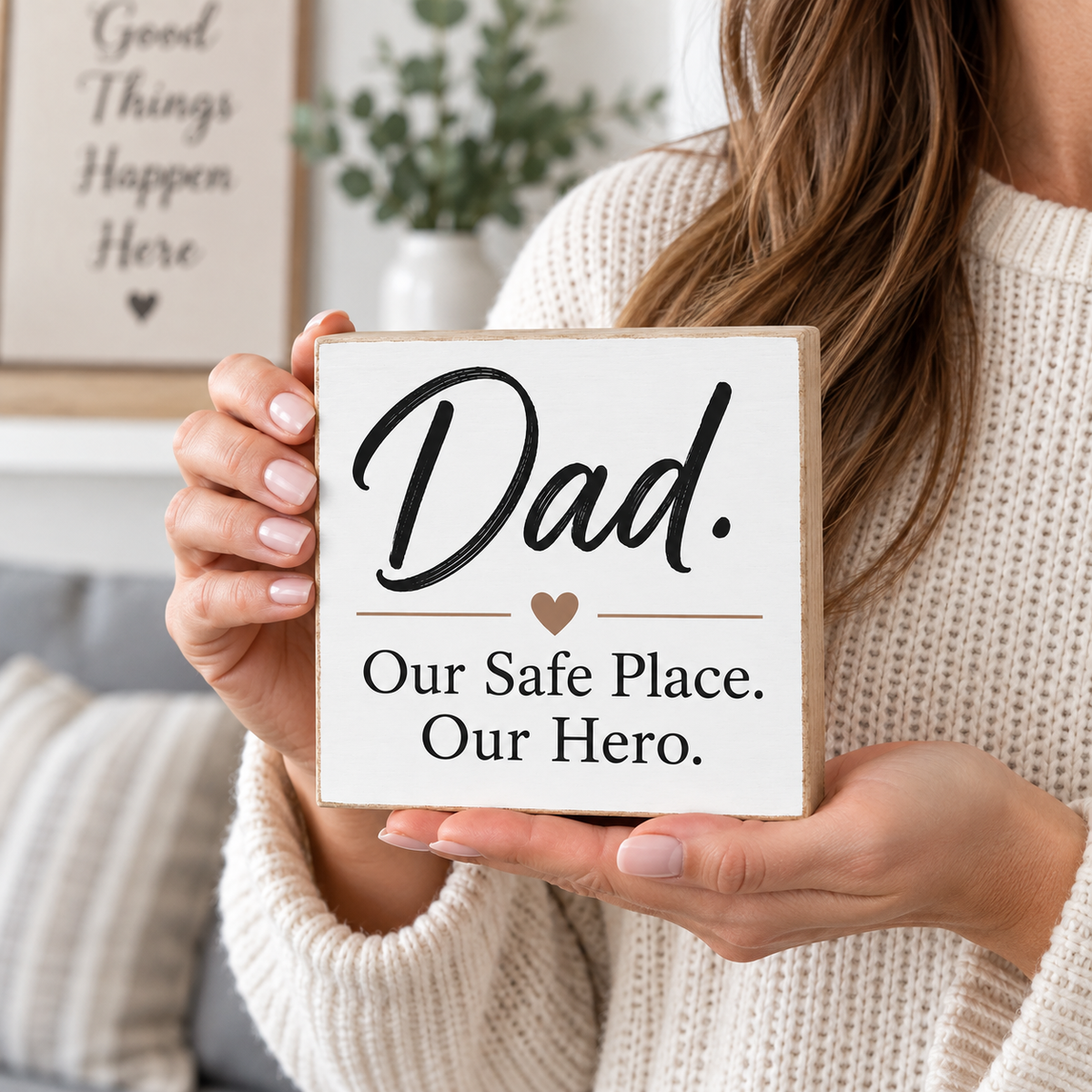 A woman in a cream knit sweater holds a square white wooden sign that reads “Dad. Our Safe Place.